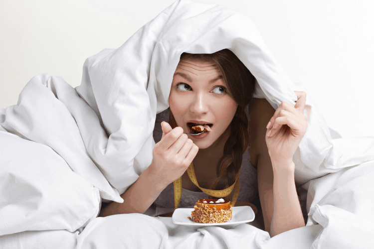 woman hiding under her covers eating dessert 